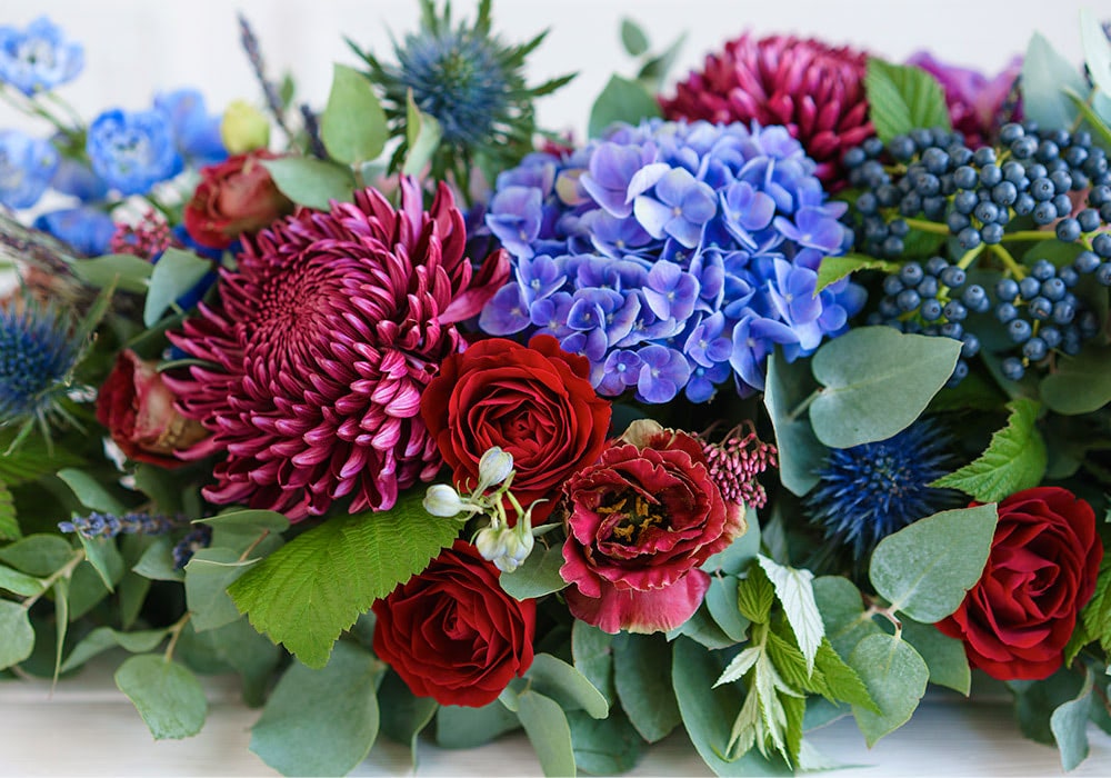 A vibrant bouquet displays red roses, purple chrysanthemums, blue hydrangeas, and green foliage arranged together on a white background.