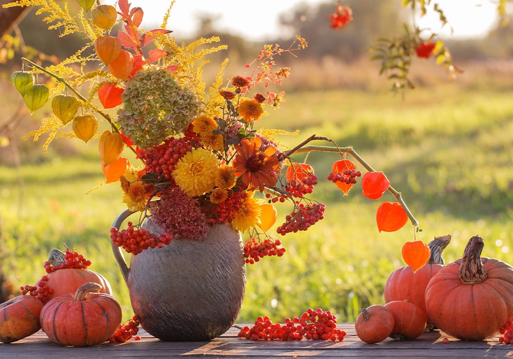 A rustic vase holds an array of colorful autumn flowers and berries, surrounded by pumpkins, set on a wooden surface outdoors with a sunlit field in the background.